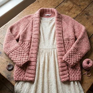 Könnte beinhalten: Ein hellrosa Cardigan mit offenem Frontdesign und strukturiertem Muster an den Ärmeln, über einem cremefarbenen Spitzenkleid. Ein Knäuel passender Wolle liegt daneben. Der Cardigan ist aus einem grob gestrickten Material.