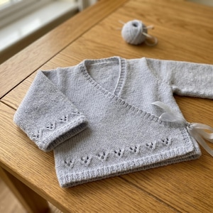 May include: A pale blue knitted baby wrap cardigan with decorative edging on the sleeves and hem. The cardigan has a wrap-around design and is tied with a white ribbon. A ball of pale blue yarn is in the background.