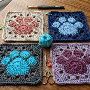 Könnte beinhalten: Vier gehäkelte Quadrate in verschiedenen Farben, jedes mit einem Pfotenabdruck-Design. Farben sind Beige, Marineblau, Türkis und Lila. Ein Knäuel türkisfarbener Wolle, ein Häkelhaken und eine Schere sind ebenfalls vorhanden.