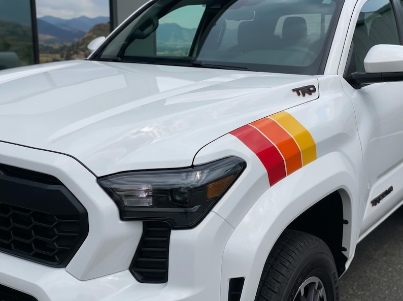 Pode incluir: Caminh&atilde;o branco com um design de listras retr&ocirc; em vermelho, laranja e amarelo. O caminh&atilde;o tem far&oacute;is pretos e as letras "TRD" s&atilde;o vis&iacute;veis no cap&ocirc;. O pneu &eacute; preto.
