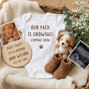 May include: A white baby onesie with the text "OUR PACK IS GROWING! COMING SOON" and a paw print. A wooden sign says "PLOT TWIST: IT'S A HUMAN! BABY WILSON EST. OCT 2026". Includes a dog photo, ultrasound image, dog plush, and brown baby booties.