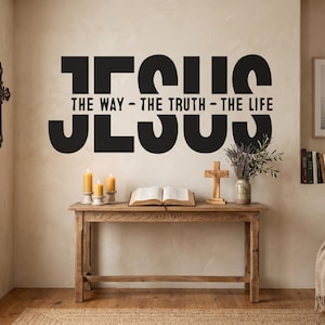 May include: Black wall art with the word "JESUS" and the phrase "THE WAY - THE TRUTH - THE LIFE". A wooden table holds candles, a book, a cross, and a vase of flowers. A framed picture and a cross are also visible.