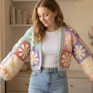 Può includere: Un cardigan colorato all'uncinetto con un motivo a margherita. La giacca presenta un mix di colori pastello, tra cui azzurro, rosa, giallo e viola. Le maniche sono lunghe e a sbuffo e il cardigan è aperto sul davanti.