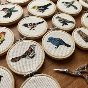 May include: A collection of small, round embroidery hoops featuring cross-stitched bird designs. The birds are depicted in various colors and poses, set against a white fabric background. A pair of silver scissors rests nearby. The hoops are arranged on a wooden surface.