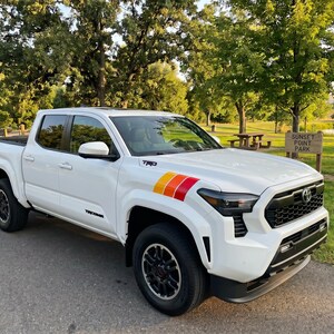 Pode incluir: Uma pick-up Toyota Tacoma branca com rodas pretas e um design de listras coloridas no cap&ocirc;. A pick-up est&aacute; estacionada em uma estrada pavimentada perto de um parque com uma placa "Sunset Point Park".