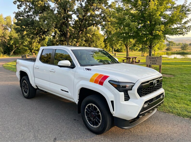 Pode incluir: Uma pick-up Toyota Tacoma branca com rodas pretas e um design de listras coloridas no cap&ocirc;. A pick-up est&aacute; estacionada em uma estrada pavimentada perto de um parque com uma placa "Sunset Point Park".