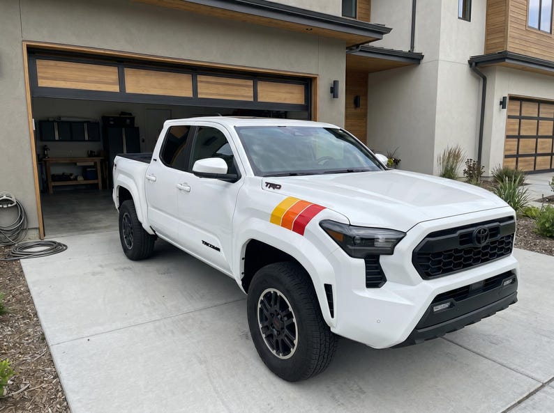 Pode incluir: Caminhonete Toyota Tacoma branca com rodas e acabamentos pretos. Possui um decalque de listras coloridas no painel lateral dianteiro. A caminhonete est&aacute; estacionada em uma entrada em frente a uma casa moderna com garagem.