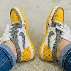 Può includere: Un paio di pantofole a forma di sneaker fatte a mano all'uncinetto. Le pantofole sono gialle, grigie e bianche, con lacci bianchi. Il design imita l'aspetto di un popolare stile di sneaker. Le pantofole sono progettate per essere indossate in casa.
