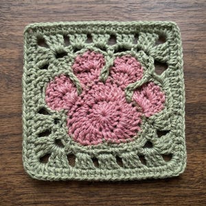 Puede incluir: Un posavasos cuadrado tejido a ganchillo con un diseño de huella de pata rosa en el centro, rodeado por un borde verde. El posavasos hecho a mano descansa sobre una superficie de madera. El diseño de la huella es encantador.