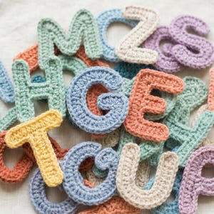 Può includere: Un insieme di lettere all'uncinetto dai colori pastello assortiti, tra cui blu, verde, arancione, giallo e viola. Le lettere sono sparse su una superficie chiara e strutturata, creando un'estetica giocosa e artigianale. Ideale per scopi educativi o artigianali.