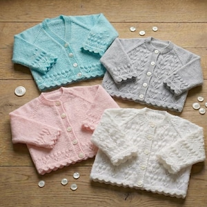 Puede incluir: Cuatro cárdigans de bebé de punto en colores pastel, azul, gris, rosa y blanco, cada uno con cierres de botón y bordes festoneados. Varios botones blancos están dispersos alrededor de las prendas sobre una superficie de madera.