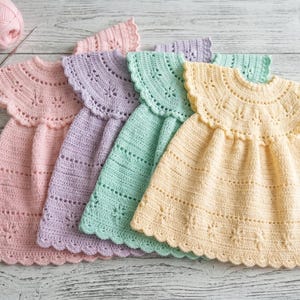 May include: Four pastel-colored crochet dresses in pink, lavender, mint green, and yellow are displayed on a light wood surface. Each dress features a scalloped neckline and a detailed floral pattern, showcasing handmade craftsmanship. A ball of pink yarn and a crochet hook are in the background.