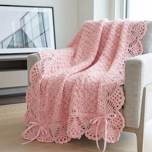 Op de afbeelding: Een lichtroze gehaakte deken gedrapeerd over een grijze fauteuil. De deken heeft een geschulpte rand en is vastgebonden met roze linten. Een ingelijste foto hangt op de achtergrond aan de muur.