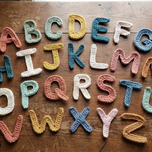 May include: A collection of colorful, crocheted letters of the alphabet arranged on a wooden surface. The letters are in various colors, including coral, teal, yellow, and white. The handmade letters are a decorative item.