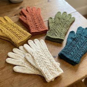 Puede incluir: Seis guantes de punto en varios colores: amarillo mostaza, naranja óxido, verde oliva, verde azulado y crema. Los guantes presentan un diseño de punto trenzado y están colocados sobre una superficie de madera.
