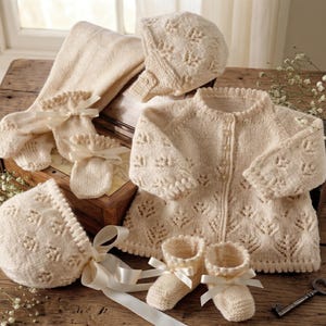 May include: A cream-colored baby clothing set, including a cardigan, bonnet, booties, mittens, and socks, all with a delicate leaf pattern. The set is tied with cream-colored ribbons and presented on a wooden surface.