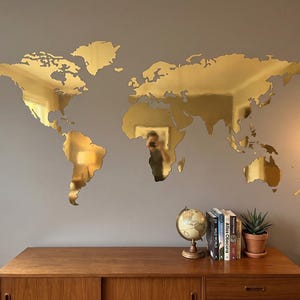 Könnte beinhalten: Eine große, goldfarbene Weltkarte aus Metall an einer grauen Wand. Die Karte zeigt die Kontinente als detaillierte Umrisse. Darunter befindet sich ein Holzschrank mit einem Globus, Büchern und einer Topfpflanze, die eine dekorative Ausstellung bilden.