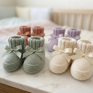 May include: Six pairs of knitted baby booties in pastel shades of sage green, cream, lavender, and peach. Each bootie has a satin ribbon bow. The booties are displayed on a white surface.