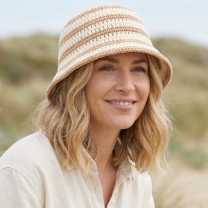 May include: A crocheted bucket hat with alternating beige and cream horizontal stripes. The hat has a slightly downward-sloping brim. The person in the image is wearing a light-colored button-down shirt.