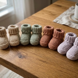 Puede incluir: Cuatro pares de patucos para bebé tejidos a mano dispuestos en fila. Los patucos presentan un puño acanalado y una puntera en punto bobo. Los colores incluyen crema, verde salvia, terracota y lavanda suave, con un acabado de lana texturizada.