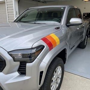 Pode incluir: Caminhonete prateada com um design de listras coloridas na frente. A caminhonete tem far&oacute;is pretos e uma grade preta. A caminhonete est&aacute; estacionada em uma garagem.