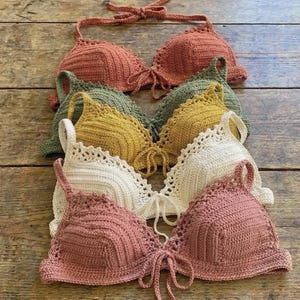 May include: A collection of handmade crochet tops in various colors, including rust, olive green, mustard yellow, cream, and dusty rose. Each top features a halter neck tie and a detailed, textured stitch pattern. The tops are arranged on a wooden surface.