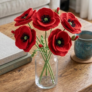 以下が含まれることがあります： 手作りの赤いかぎ針編みのポピーの花束。黒い中心と緑の茎があり、透明なガラスの花瓶に入っています。木製のテーブルの上にあり、青いマグカップと本が近くにあります。