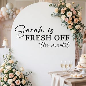 May include: A white arched backdrop featuring the black text "Sarah is FRESH OFF the market" in a mix of script and serif fonts. The display is decorated with arrangements of cream and blush roses and green eucalyptus foliage.