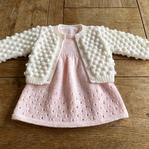 Pode incluir: Um conjunto de malha para bebé, com um vestido rosa claro com um delicado padrão de ilhós e um cardigan creme. O cardigan tem um design texturizado de ponto de arroz. O conjunto é apresentado numa superfície de madeira.