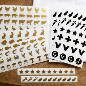 May include: Sheets of gold and black stickers featuring various designs. Gold stickers include animal and food shapes like chickens, pigs, cows, ducks, rabbits, fish, crabs, and acorns. Black stickers have stars, crosses, and vegetables.