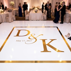 May include: A white dance floor with gold trim and large gold initials "D & K" and the date "May 17, 2027". The floor is in a ballroom with tables and guests in the background. A pair of shoes are on the floor.