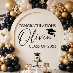 May include: A graduation party scene with a large arch decorated with gold, black, and white balloons. The arch features the text "Congratulations Olivia Class of 2026" and a graduation cap graphic. A cake and desserts are on display.