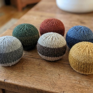 Può includere: Sei sfere lavorate a mano a maglia in vari colori: grigio, verde, ruggine, blu, beige con una striscia marrone e giallo. Gli oggetti sono disposti su un tavolo di legno, mostrando le loro superfici lavorate a maglia.