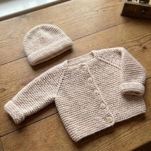 Könnte beinhalten: Hellbeige gestricktes Baby-Set, bestehend aus einer Strickjacke mit fünf Knöpfen und einer passenden Mütze. Das Set ist aus einem grob gestrickten Stoff gefertigt und wird auf einer Holzoberfläche präsentiert. Die Jacke hat lange Ärmel und gerippte Bündchen.