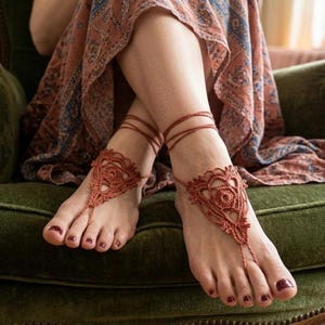 Könnte beinhalten: Rostfarbene Häkel-Barfußsandalen mit aufwendigen floralen Designs. Die Sandalen verfügen über eine Zehenschlaufe und Knöchelbänder, die einen zarten und böhmischen Look erzeugen. Das Bild zeigt die Sandalen an den Füßen einer Person.