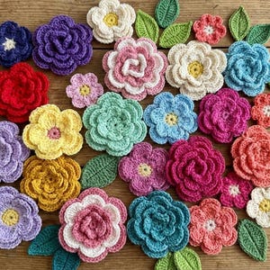 Könnte beinhalten: Eine Sammlung bunter, gehäkelter Blumen in verschiedenen Größen und Farbtönen, darunter Rot, Lila, Gelb, Rosa und Blau. Grüne gehäkelte Blätter sind zwischen den Blumen verstreut, alles auf einer Holzoberfläche angeordnet.