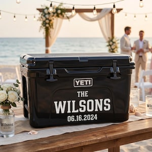 May include: A black YETI cooler with the text "THE WILSONS 06.16.2024" in white, set on a wooden table at a beach wedding. The scene includes floral arrangements, glasses, and a beach backdrop.