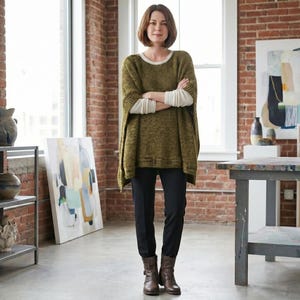 Pode incluir: Uma mulher num estúdio de arte com um suéter verde, uma camisa branca de manga comprida, calças pretas e botas castanhas. O estúdio apresenta obras de arte, cerâmica e uma grande mesa de madeira. As paredes são de tijolo.