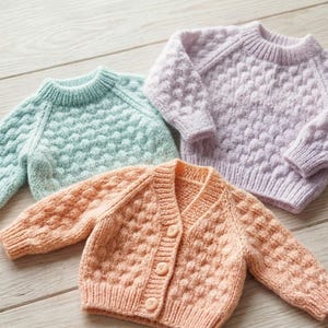 Puede incluir: Tres suéteres de bebé tejidos en colores pastel: azul claro, melocotón y lavanda. Los suéteres tienen un patrón texturizado, similar a burbujas, y mangas largas. Uno es un cárdigan con botones.