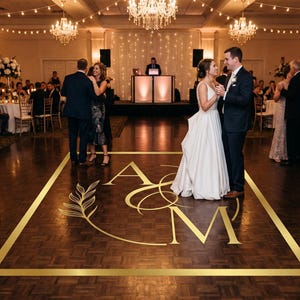 May include: A wedding reception with a dance floor featuring a gold geometric design with the initials "A" and "M". A bride and groom dance in the center, surrounded by guests and chandeliers. The floor is a dark wood.