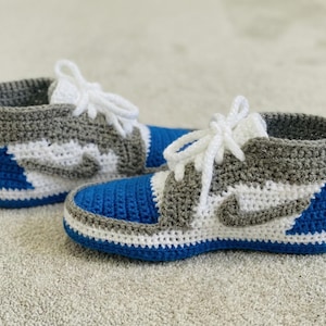 Könnte beinhalten: Ein Paar handgehäkelte Sneaker in Blau, Weiß und Grau. Die Sneaker haben ein klassisches High-Top-Design mit weißen Schnürsenkeln und einem grauen Nike-Swoosh. Zehen und Ferse sind blau, mit einem weißen Streifen.