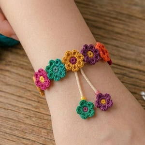 Pode incluir: Uma pulseira de crochê colorida com desenhos de flores em tons de rosa, verde-azulado, laranja e roxo. A pulseira é adornada com pingentes de flores penduradas. A imagem também mostra novelos de linha e uma agulha de crochê.