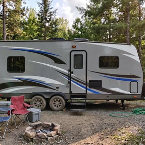 Puede incluir: Una caravana gris con detalles decorativos azules y negros está estacionada en un camping arbolado. Dos sillas plegables, una nevera portátil y una hoguera están frente a la caravana. Una manguera verde está enrollada cerca.