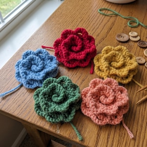 Könnte beinhalten: Fünf gehäkelte Blumenapplikationen in Blau, Rot, Gelb, Grün und Pfirsich liegen auf einer Holzoberfläche. Stricknadeln, Knöpfe und Garn sind ebenfalls zu sehen. Die Blumen haben ein Spiraldesign und einen Stiel.