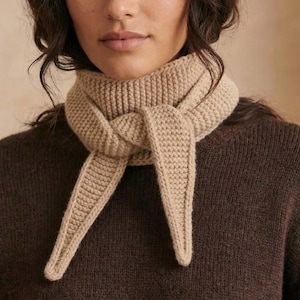 May include: A beige knitted scarf tied in a knot, worn over a brown sweater. The scarf has a thick, textured appearance, with the ends hanging down. The sweater has a simple, classic design.