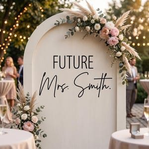 May include: A cream-colored wooden sign with an arched top, adorned with floral arrangements and the text "FUTURE Mrs. Smith." The sign is decorated with pampas grass, roses, and greenery, perfect for a wedding or engagement celebration.