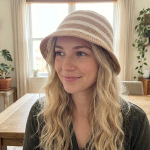 May include: A beige and white striped bucket hat. The hat has a rounded crown and a short brim. The hat is made of a woven material. The hat is worn by a person with long, wavy blonde hair.