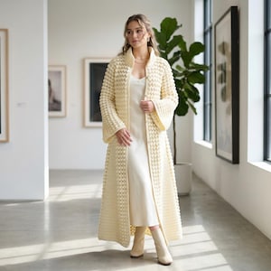 Könnte beinhalten: Ein langer, cremefarbener Mantel mit offenem Vorderteil und einem strukturierten, blasenartigen Muster. Der Mantel hat weite Ärmel und wird über einem weißen Kleid getragen. Das Outfit wird mit cremefarbenen Stiefeln kombiniert. Der Hintergrund ist ein gut beleuchteter Raum mit Kunstwerken.