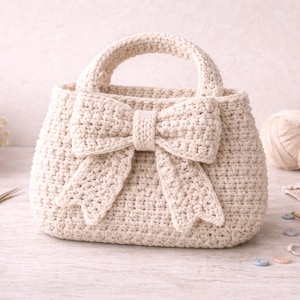 May include: Cream-colored crochet handbag with a large bow detail on the front. The bag has a structured shape with a top handle. The background includes yarn, scissors, and other crafting supplies, suggesting a handmade item.