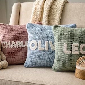 Könnte beinhalten: Drei Häkelkissen in Rosa, Blau und Grün, jedes mit einem Namen in weißen Buchstaben: CHARLO, OLIV und LEO. Ein Stoffhund mit Fliege und ein Holzspielzeug in Regenbogenfarben sind ebenfalls zu sehen.